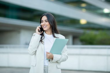 Beyaz ceketli ve beyaz gömlekli bir iş kadını bir ofis binasının önünde duruyor. Telefonuyla konuşuyor ve elinde mavi bir dosya tutuyor. Kopya alanı.