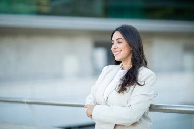 Beyaz ceketli bir kadın kollarını kavuşturup korkuluklara yaslanıyor. Uzun siyah saçları var, ve yan tarafa bakarken gülümsüyor, boşluğu kopyalıyor.