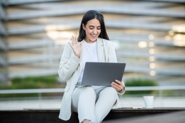 Beyaz takım elbiseli genç bir kadın modern bir ofis binasının önündeki bankta oturuyor. Bir dizüstü bilgisayar kullanıyor ve kameraya el sallıyor. Bir video çağrısına katıldığını gösteriyor.