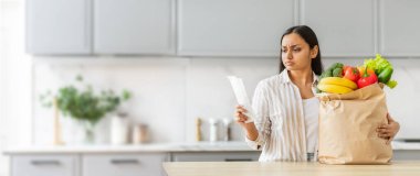 Hintli bir kadın mutfağında elinde şok olmuş bir ifadeyle fişe bakıyor. Elinde içi yeni mallarla dolu bir kese kağıdı var.