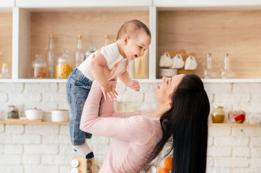 Bir kadın, hem gülümseyen hem de birbirine bakan bir bebeği sevinçle havaya kaldırır. Bebek kolları açılır ve ikisi arasında mutluluk ve bağ kurulur..