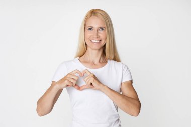 Sarı saçlı bir kadın beyaz gömlek giyiyor ve gülümsüyor. O, elleri göğsünün önünde bir kalp şekli yapıyor. Arka plan düz beyaz bir duvar..