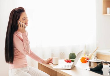 Mutfak tezgahında oturan bir kadın cep telefonuyla konuşuyor. Odaklanmış ve özenli görünüyor, muhtemelen önemli konuları tartışıyor..