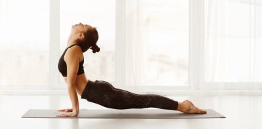Esnek kadın, iyi aydınlatılmış bir yoga stüdyosunda yukarı bakan köpek pozu veriyor. Doğal ışık odayı doldururken duruşuna odaklanmış, sakin ve huzurlu bir atmosfer yaratıyor, panorama.