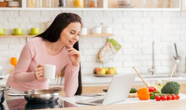 Bir kadın mutfak tezgahında oturuyor, önünde dizüstü bilgisayarla meşgul. İnternette gezinirken ya da bir proje üzerinde çalışırken odaklanmış ve konsantre olmuş görünüyor..