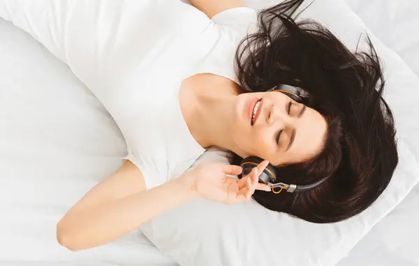 stock image A woman in a white shirt lies on her bed, enjoying music through headphones. She looks relaxed and happy, indicating a peaceful and enjoyable moment.