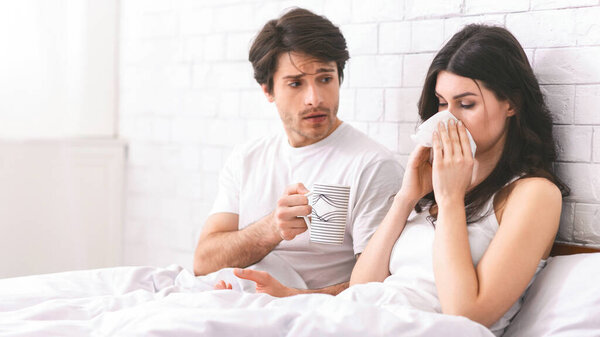 A man with a worried expression holds a mug of tea while a sick woman blows her nose with a tissue in bed. The couple are in a bedroom with a white brick wall.
