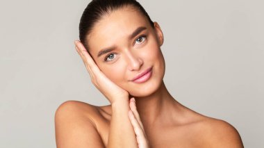 Bir portre için poz veren uzun saçlı güzel bir genç kadın. O, yanaklarına karşı ellerini tutuyor, özelliklerini vurguluyor. Sakin bir ifadeyle doğrudan kameraya bakıyor..