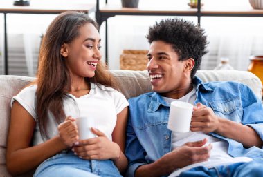 Neşeli siyah genç çift kanepede birlikte oturur, gülümserler ve kahve kupaları tutarlar. Rahatlamış ve mutlu görünüyorlar, birbirlerine eşlik etmekten zevk alıyorlar..