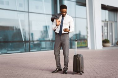 Afro-Amerikalı iş adamı, yanında bavullarıyla modern bir havaalanının girişinde bir telefon görüşmesi yapıyor. Ortam profesyonel ve hareketli, seyahat ve iletişimi gösteriyor..