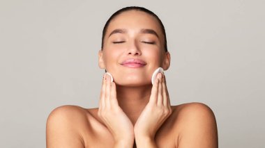 Genç bir kadın stüdyo ortamında pamuk pedlerle nazikçe cilt ürünleri uygularken görülüyor. Gözleri kapalı ve kendine bakmanın keyfini çıkarıyor gibi görünüyor..