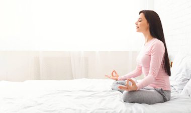 Genç bir kadın bir yatakta nilüfer pozisyonunda oturur ve huzur içinde meditasyon yapar. Rahat kıyafetler giyer ve beyaz perdelerin arasından güneş ışığı filtreleri geçirir..