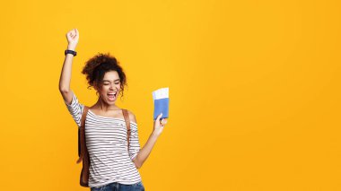Kıvırcık saçlı Afro-Amerikalı genç bir kadın zafer için elini kaldırırken heyecan saçıyor. Elinde bir pasaport ve sarı arka planda biletler var. Kopya alanı.