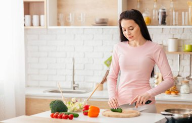 Bir kadın ahşap kesme tahtasıyla sebze doğramakla meşgul. Etrafında havuç, dolma biber ve soğan gibi çeşitli sebzeler var..