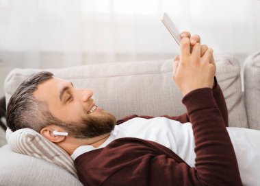 Kulaklıklı sakallı bir adam, iyi aydınlatılmış bir oturma odasında akıllı telefon kullanırken koltukta rahat bir şekilde uzanır ve gülümser. Rahat duruşu, bir anlık boş zamanın tadını çıkardığını gösteriyor..