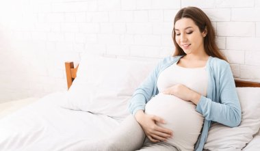 Hamile bir kadın bir yatakta oturuyor. O, elleri yavaşça bebek yumrusunu sarkıtırken, rahatça dinleniyor. Oda hafifçe aydınlatılmış, huzurlu bir atmosfer yaratıyor..