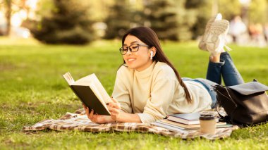 Asyalı kadın bir kitap okumaya dalmış, çimlerin üzerinde uzanıyor. Doğayla iç içe, arka planda ağaçlar ve çiçekler var. Güneş parlıyor, sahne üzerine sıcak bir parıltı saçıyor..
