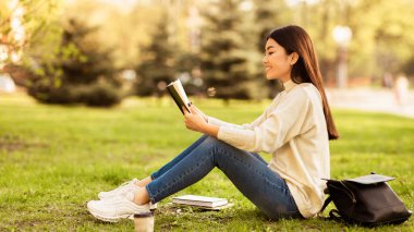 Asyalı bir kadın, okuduğu bir kitaba dalmış çimlerin üzerinde oturuyor. Güneş ışığı, açık havada okumaktan ve yandan bakmaktan hoşlanırken onun özelliklerini vurguluyor.