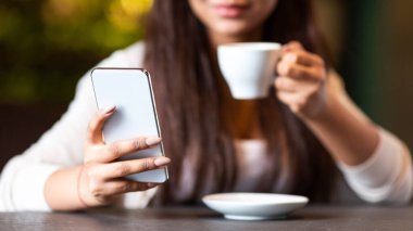 Kırpılmış kız bir elinde bir fincan kahve, diğer elinde bir cep telefonu tutarken oturuyor. İçkisini yudumlarken telefonla meşgul gibi görünüyor..