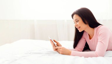 Elindeki cep telefonu ekranına odaklanmış bir kadın yatakta yatıyor. Cihaza kendini kaptırmış görünüyor, rahat bir duruşu var. Oda loş ışıkta, ekranların parlaklığını vurguluyor..