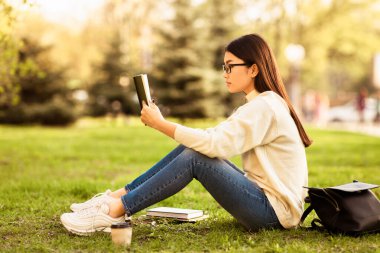 Asyalı genç bir kadın, bir kitap okumaya dalmış, sakin bir parkta çimlerin üzerinde oturuyor. Etrafı ağaçlarla çevrili, sıcak, güneşli havanın tadını çıkarıyor yanında buzlu bir kahve ve yanında bir sırt çantası ile..