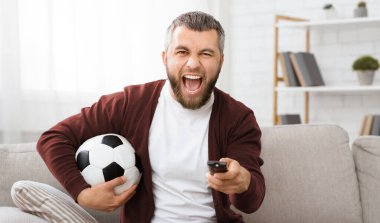 Bir elinde futbol topu, diğer elinde uzaktan kumanda olan bir adam görülüyor. Görünüşe göre birden fazla iş yapıyor ya da televizyon izlemekle futbol oynamak arasında geçiş yapıyor..