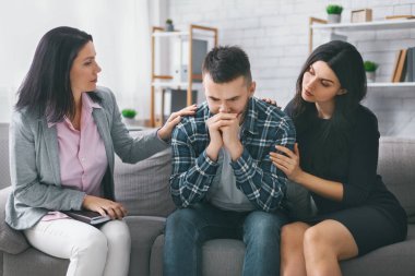 Partneri ve terapisti ile koltukta oturan bir adam. İkisi de ellerini omzuna koyup destek oluyor. Zor bir duygusal an yaşıyor.