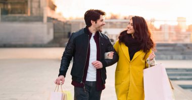 Bir adam ve bir kadın alışveriş torbaları tutarken binanın önünde dikiliyorlar..