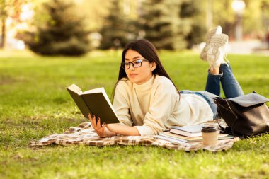 Asyalı bir kadın okuduğu bir kitaba dalmış çimlerin üzerinde uzanıyor. Edebiyat dünyasına dalmış sayfaları çevirirken güneş tepemizde parlıyor..