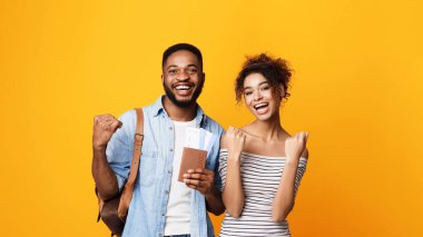 Afro-Amerikalı neşeli genç adam ve kadın yanyana durur, mutluluk ve enerji saçarak canlı sarı bir zemin üzerinde, pasaportlar ve jestler tutarak
