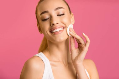 Makyaj çıkarıcıyı yanağına pamukla sürerken gülümseyen genç bir kadın görülüyor. Arka plan sağlam, parlak bir pembe.