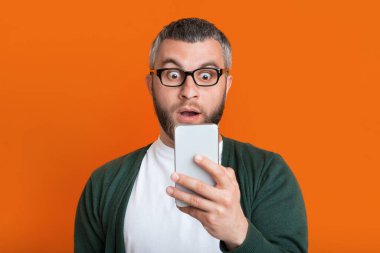 Gözlüklü, yeşil hırka ve beyaz gömlek giyen sakallı bir adam, akıllı telefonundaki bir mesajı turuncu bir arka planda okurken şaşırmış görünüyor..