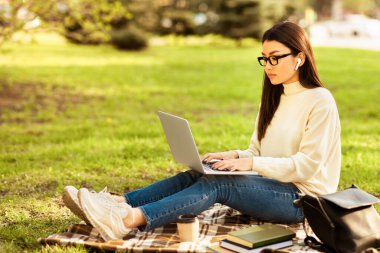 Asyalı bir kadın dışarıda bir battaniyenin üzerinde oturuyor, dizüstü bilgisayarda yazıyor. Açık bir alanla çevrili olan ekrana odaklanmış durumda. Güneş parlıyor ve arka planda ağaçlar var..