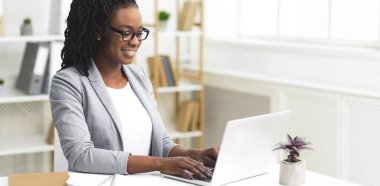 Gülümseyen Afro-Amerikan iş kadını bilgisayarını masasında kullanıyor. Çağdaş ofis ortamında, içinde fotokopi alanı olan panorama.