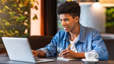 Genç bir Afro-Amerikalı bir adam bir kafede oturuyor ve dizüstü bilgisayarına odaklanıyor. Kahve fincanları ve defterlerle çevrili klavyeye basıyor ve tıklıyor..