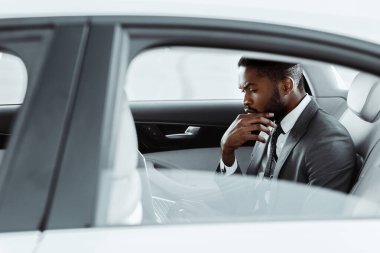 Sakallı, takım elbiseli ve kravatlı Afrikalı Amerikalı düşünceli bir işadamı modern bir sedanın arka koltuğunda oturuyor. Eli, çenesinde duruyor ve bakışları yere eğik.