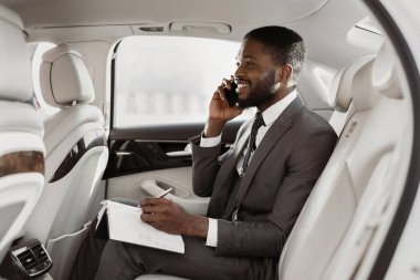 Siyah işadamı bir arabanın arka koltuğunda oturuyor, telefonla konuşurken bir not defterine not alıyor. Takım elbise ve kravat takıyor ve yüzünde ciddi bir ifade var..