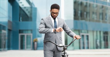 Bisikletli profesyonel bir siyah adam şehir merkezinde saatine bakıyor. Sahne modern ve enerjik, profesyonel sorumlulukların ve işe gidip gelişin karışımını gösteriyor..
