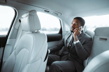 Gri takım elbiseli Afro-Amerikalı işadamı bir arabanın arka koltuğunda oturuyor ve cep telefonuyla konuşuyor. O, yüzünde düşünceli bir ifadeyle pencereden dışarı bakıyor..