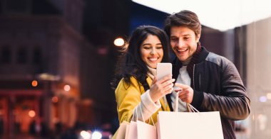 Bir çift, telefon ekranlarına bakarken ellerinde alışveriş torbalarıyla dışarıda dikiliyor..
