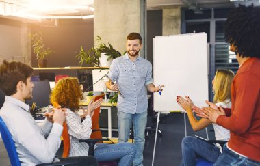 Bir adam bir grup özenli bireyin önünde duruyor, bir sunum sunarken güvenle el kol hareketi yapıyor ve konuşuyor. Seyirci oturuyor, dikkatle dinliyor, not alıyor.