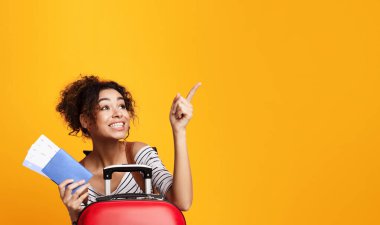 Afro-Amerikalı bir kadın sarıya basıyor, bir elinde kırmızı bir bavul tutuyor ve fotokopi alanını işaret ediyor. Kamera dışında bir şeye doğru hareket ederken odaklanmış ve kararlı görünüyor..