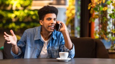 Genç siyahi bir adam masada oturuyor ve bir telefon görüşmesi yapıyor. Telefonda konuşurken odaklanmış görünüyor..