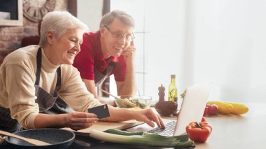 Yaşlı bir çift modern bir mutfakta birlikte yemek pişiriyor. Dizüstü bilgisayar ve kredi kartı, market siparişi, kopyalama alanı