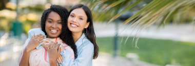 Açık havada sarılıp poz veren iki çok ırklı kız arkadaşın portresi. Kameraya sarılıp gülümseyen güzel genç bayanlar.