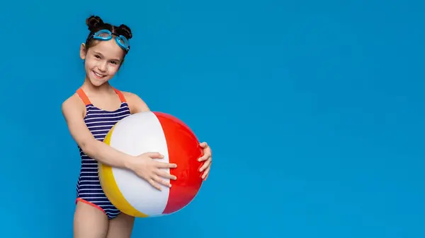 stock image Beach entertainment. Little girl in swimwear smiling and holding big inflatable ball, blue studio background