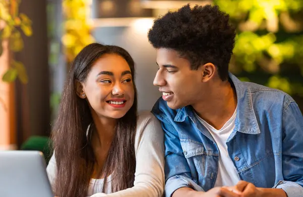 stock image A young black guy and girl sitting at a table in a cafe, both focused on the screen of a laptop in front of them. They appear to be engrossed in whatever is displayed on the screen