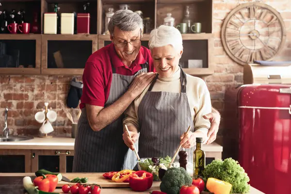 Kısa, gri saçlı, beyaz kazaklı ve gri çizgili önlüklü son sınıf öğrencisi bir kadın, duvarlı bir mutfakta salata hazırlıyor. Bir kase yeşili tahta kaşıkla karıştırırken gülümsüyor.