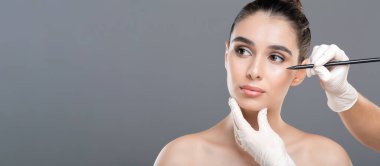 Genç bir kadın bir klinikte kozmetik tedavi görüyor. Bir profesyonel yüzünü kalemle işaretlerken diğer el çenesini tutuyor..