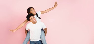 Neşeli bir zenci, pembe arka planda gülümseyen bir kızı sırtında taşır. Her ikisi de günlük beyaz tişörtler ve kot pantolon giyiyor.